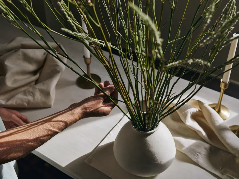 Mâini aranjând tulpini uscate într-un vas ceramic, pe un blat de lucru